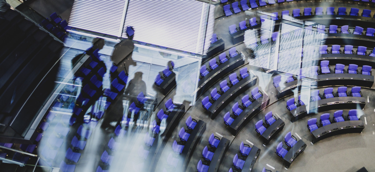 Ein Bild des Bundestag-Plenarsaals, schemenhaft sind Menschen in einer Spiegelung erkennbar.