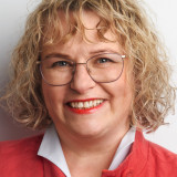 Portraitfoto einer blonden, lächelnden Frau mit welligem, mittellangem Haar, schmal eingefasster großer Brille, die eine weiße Bluse und einen knallroten Mantel trägt. 