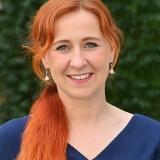 Portrait von Franziska Schubert im Sommer mit einem Lächeln in einem dunkelblauen Kleid vor dunkelgrünem Pflanzenhintergrund.