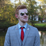 Kompetenter junger Politiker in einem grauen Anzug mit blauem Hemd, roter Krawatte und stylischer Sonnenbrille vor einem idyllischen grünen Hintergrund.