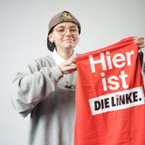 Bild von Kandidatin "Loo" Elena Krein mit roter Weste in der Hand auf der "Hier ist DIE LINKE" steht