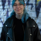 Ein Portrait einer femininen Person mit schulterlangen, blauen Haaren. Sie trägt eine Brille, hat Piercings und eine Motte auf dem Hals tätowiert. Sie trägt ein schwarzes Oberteil und eine schwarze Lederjacke und steht vor einer Wand mit Graffiti, die unscharf im Hintergrund zu sehen ist.