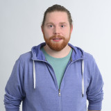 Simon Metzger, ein junger Mann mit rotem Bart steht vor neutralem Hintergrund. Er trägt einen lila-farbene Jacke