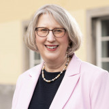 Portraitbild von Ulla Weiß vor dem Rathaus in Witten. Ulla trägt ein rosa Jackett, ein blaues T-Shirt mit Halskette und lächelt freundlich in die Kamera.