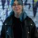 Ein Portrait einer femininen Person mit schulterlangen, blauen Haaren. Sie trägt eine Brille, hat Piercings und eine Motte auf dem Hals tätowiert. Sie trägt ein schwarzes Oberteil und eine schwarze Lederjacke und steht vor einer Wand mit Graffiti, die unscharf im Hintergrund zu sehen ist.